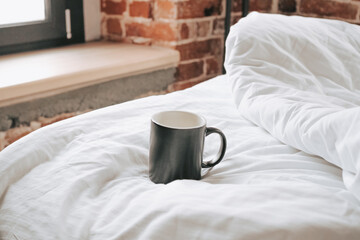 Fototapeta na wymiar Black mug with hot coffee on a white blanket in the morning. Early wake-up and morning coffee in the hotel bed. Stylish apartments and white bed linen