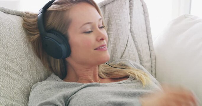 Listening to my last e-book on pregnancy. Beautiful young pregnant woman relaxing on sofa and listening to podcasts. Happy mother to be using headphones and digital tablet, browsing online, technology