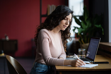 Young businesswoman prepare for financial report presentation study sales data make notes in notebook. Concentrated finance specialist writing ideas for business strategy development looking at laptop