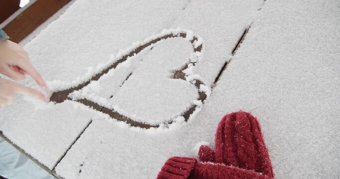 Young girl drawing heart on wooden table covered with snow outdoors, closeup