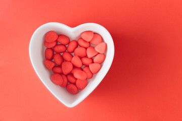 Red candies in a white heart-shaped bowl on a red background. Minimal Valentine's Day flat lay concept