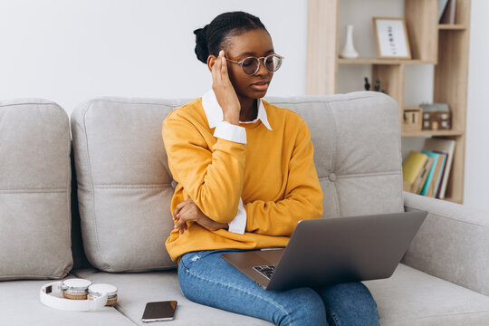 Smiling Young Black Woman In Yellow Sweater Freelancer Working From Home, Sitting On Sofa, Using Laptop, Copy Space And Thinking Seriously.