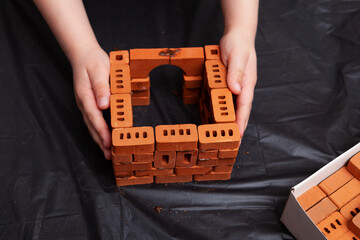 Children's hands build toy house using real clay bricks, roof tiles and cement