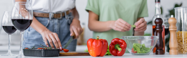cropped view of lesbian couple cooking salad near glasses of red wine in kitchen, banner.