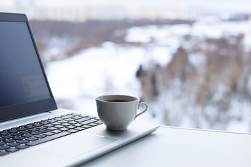 Laptop with coffee cup on a table against the window, view to winter city and park. Cozy workplace in home office