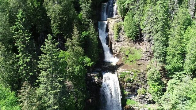 A Multiple Step Waterfall Is In View Between Tall Green Trees. This aerial view slowly backs away from the main subject throughout the video clip.