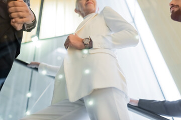 Fototapeta premium low angle view of senior businesswoman in white clothes near interracial bodyguards on hotel escalator.