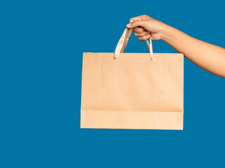 Right hand holding a brown paper bag with handle isolated on blue background.