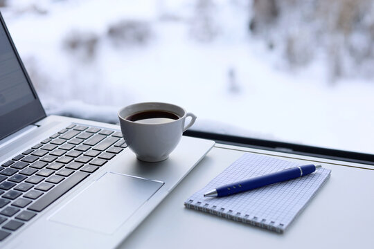 Laptop, coffee cup and notepad with pen on a table against the window, view to winter city. Cozy workplace in home office