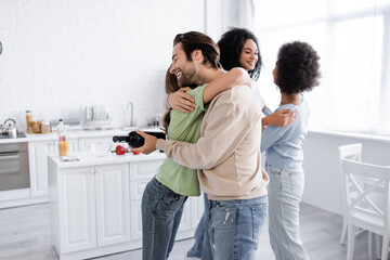 happy man with bottle of wine hugging woman near happy african american friends at home.