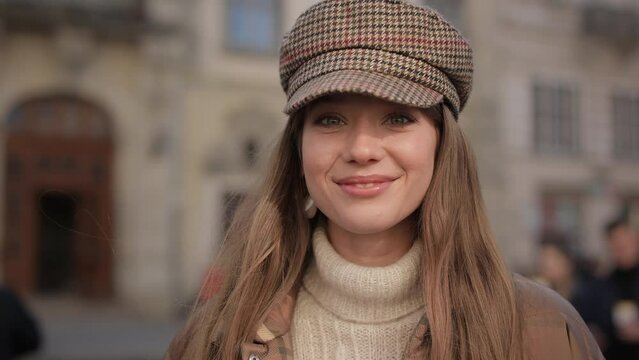 Portrait of charming caucasian woman wearing trendy hat and jacket standing on city street and looking at camera. Pretty female face.