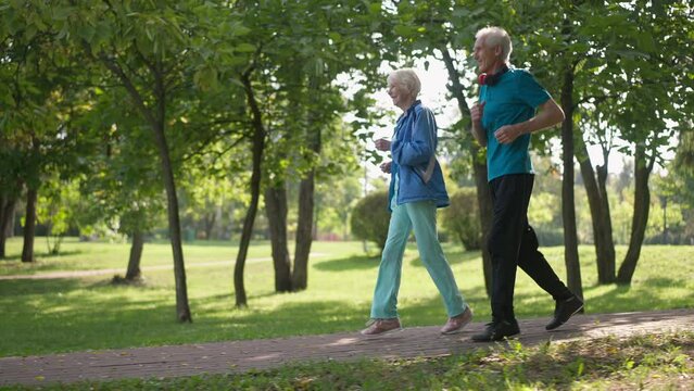 Side view happy senior couple running in slow motion in sunrays outdoors smiling. Wide shot confident Caucasian fit man and woman jogging in sunshine in spring summer park. Sport and love