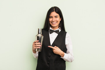young pretty hispanic woman smiling cheerfully, feeling happy and pointing to the side. cocktail bartender concept - obrazy, fototapety, plakaty