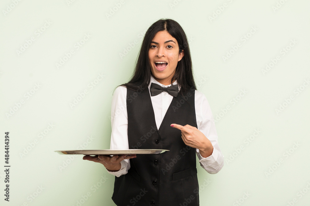 Wall mural young pretty hispanic woman looking excited and surprised pointing to the side. waiter with tray con