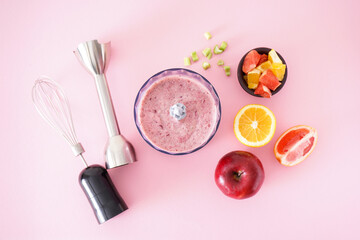 Black plastic electric hand blender and accessories with fruits and smoothie ready on pink background. Flat lay.