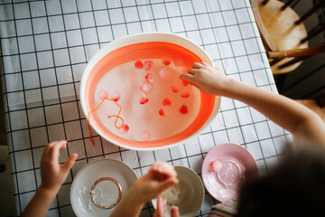Funny children play with hearts and water at home in the kitchen. Educational classes and experiments with children at home. Development of shallow motility and sensory development