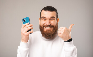 Close up portrait of a handsome bearded man showing thumb up and holding a phone. Photo over grey background.