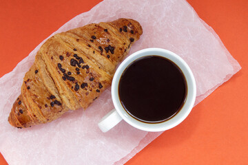 Black coffee without milk in a white cup and a chocolate croissant on parchment and bright background. French breakfast with fresh pastries. Top view flat with copy space for your text.
