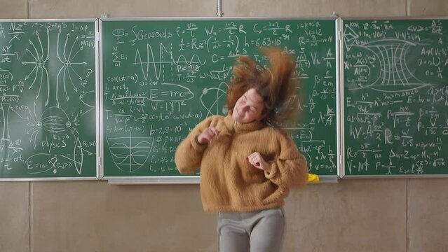 Happy female student dancing in a classroom at school against the background of a blackboard with mathematical formulas
