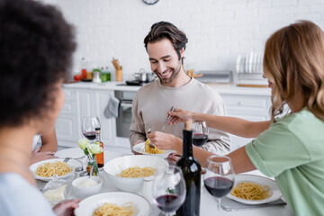 blonde woman serving pasta to cheerful man near blurred african american friend.