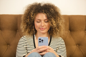 Beautiful young woman with curly hair listening to music and typing message on socialmedia app in modern blue smart phone. Attractive white female enjoying music in headphones and using mobile phone