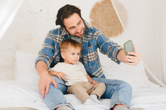 Young caucasian father dad taking selfie photo having videocall with small kid child toddler newborn infant baby at home on the bed. Videocall conversation online with relatives