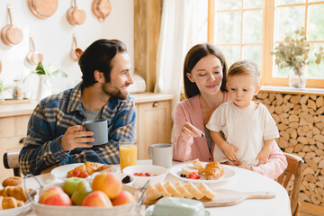 Morning breakfast at home. Young caucasian family of three, parents and infant toddler new born baby eating together, feeding son daughter at home kitchen. Parenthood concept