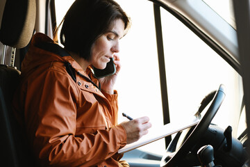 Woman talking on smartphone and writing notes on clipboard in truck