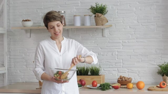 A female blogger holds a glass bowl with vegetable salad in her hands, shoots a video tutorial on cooking, a young woman at home in the kitchen tells a recipe on camera