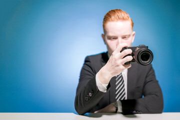 Portrait of a young red-haired man in a suit, sitting at a table taking a photo with a camera on a blue background. An office worker. Photographer