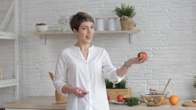 A woman holding a knife and an apple takes a video tutorial on cooking, a young woman at home in the kitchen tells the recipe on camera