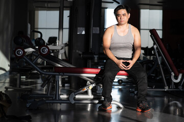 Fototapeta na wymiar i am very boredom in this place, short hair asian man look strong wear grey tank top black plant orange snaker sitting on exercise equipment get boring during workout