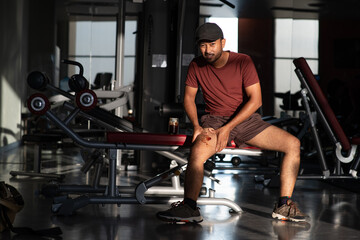 when it get hurt, black asian indian man painful face wear black cap red shirt short plant and sneaker being massaged on his leg sitting on exercise equipment during workout