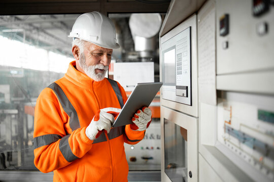 An experienced factory worker or engineer in high visibility jacket and hardhat standing by automated machine in industrial production hall using tablet computer. Factory interior.