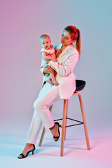A young woman in a light suit is sitting in a chair holding a baby in her arms. A businesswoman with a baby in a mixed neon pink and blue light.
