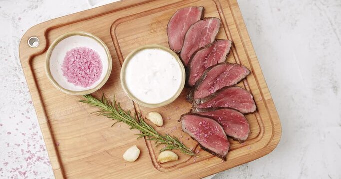 Close Up Sliced Beef Meat. Rare medium cooked beef with garlic and rosemary, white sauce and salt in a Woden plate. The salt falls on the meat. Restaurant Food. Rotating Plate on white background