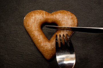 Cutting heart shape cookie with fork and knife on black stone plate. Love valentine concept. Flat lay