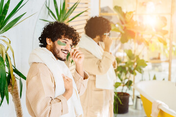 Obraz na płótnie Canvas Charismatic hispanic man in beige bathrobe enjoy morning routine in cozy bathroom interior with mirror and green plants.
