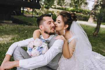 A beautiful bride in a dress hugs her back, kisses a stylish, bearded, smiling groom in a gray suit and white shirt, sitting on the green grass. Wedding portrait, photo of newlyweds.