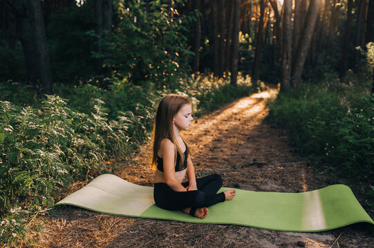 A small, hardworking, beautiful preschool girl, a child athlete sits on a green rug in a lotus position, crossing her legs, performing sports exercises, yoga and gymnastics in the forest at sunset.