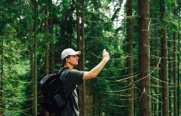 A young tourist in casual clothes and with a backpack catches the Internet in the mountains, stands in the woods with a smartphone raised and looks away. Tourism in the mountains concept.