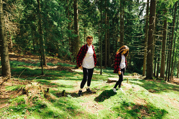 Obraz premium Beautiful man and woman standing in a sunny forest on the ground with moss and resting on a hike. Positive tourists in red shirts walk in the mountain coniferous forest.