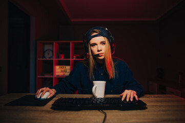 Fototapeta na wymiar Tired female gamer streaming online games on the computer at home in a cozy room, looking at the camera with a gloomy face and a cup of tea on the table, wearing a headset and cap.