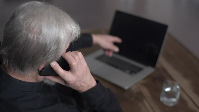 Close-up view from shoulder of unrecognizable gray-haired aged male talking to tech support and asking for help repairing broken laptop computer. User hitting keys of broken computer with fingers.