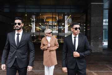 multiethnic bodyguards in sunglasses escorting mature businesswoman near hotel entrance.