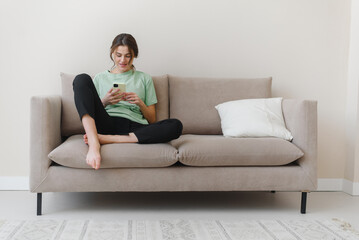 Image of happy optimistic young woman sitting indoors at home and shopping or social media using mobile phone on sofa at home.