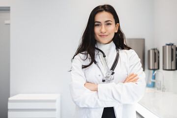Portrait of cheerful female doctor in lab coat standing with crossed hands on her office