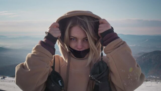 Portrait of a lovely girl enjoying ski season vacations. Blond hair moving in the wind in slow mo. Dressed in a hoodie, she fixes her hair with her hand. Mountain peaks in the snow. Winter travel.