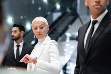 mature businesswoman checking time on wristwatch near interracial bodyguards in hotel.