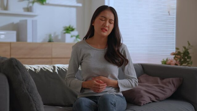 Charming asian woman stressed stomachache embracing belly sitting on sofa, Suffering from stomachache. Having menstrual painful feelings, Gastritis, Food poisoning, Abdominal or period pain concept.